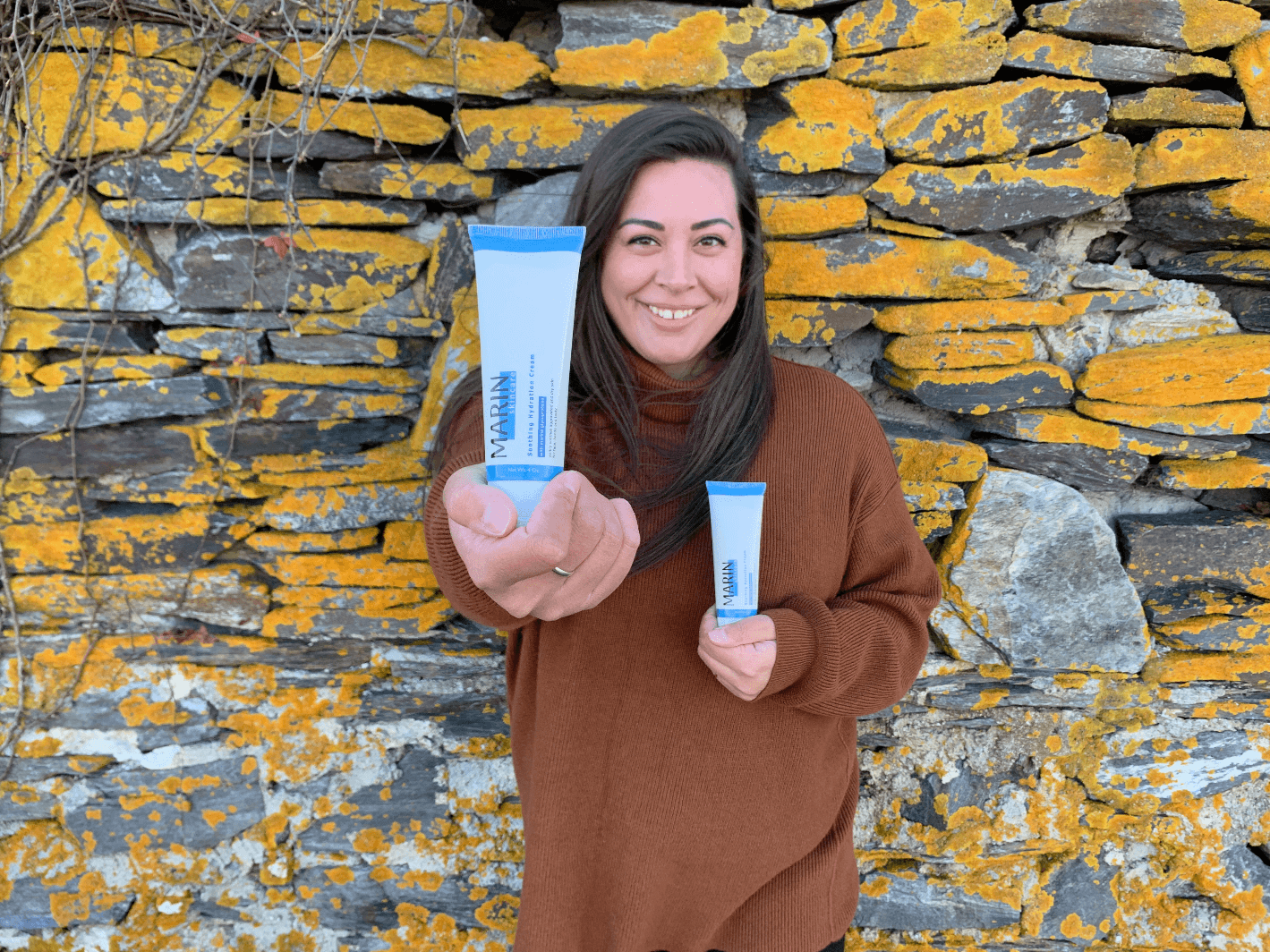 Woman smiling in brown sweater holding two MARIN skincare tubes before a lichen-covered stone wall