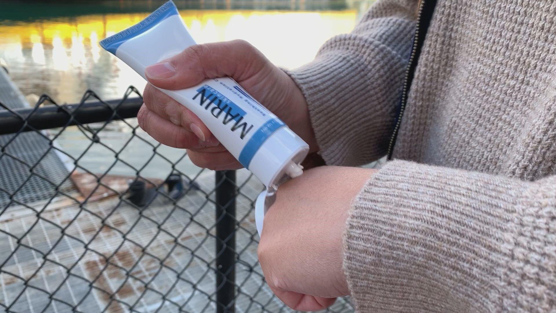 Hands squeezing a MARIN skincare tube, dispensing lotion onto the back of a hand in front of a dock and water.