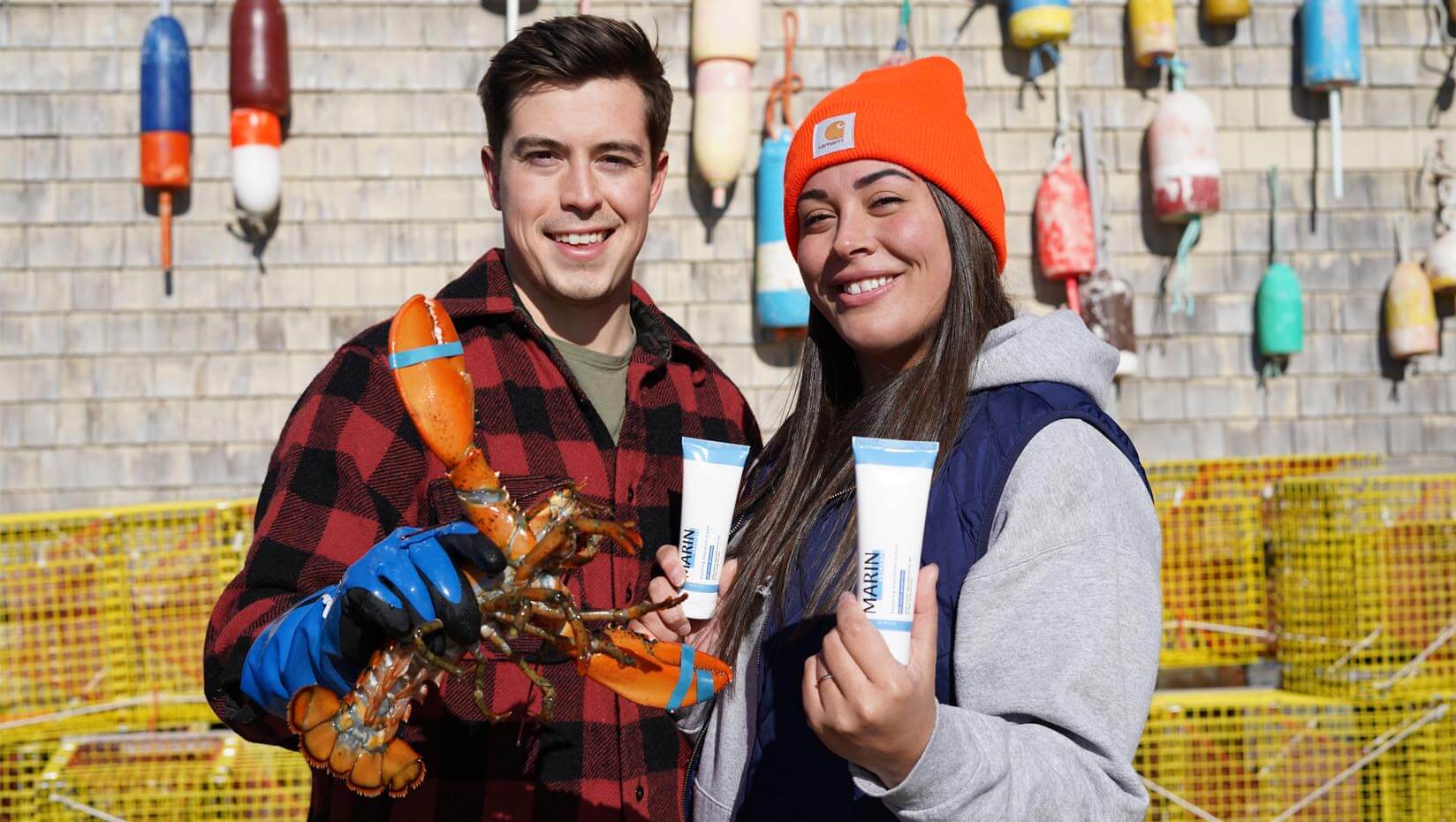 Two founders on a wharf holding a lobster and MARIN skincare tubes, smiling in front of lobster buoys