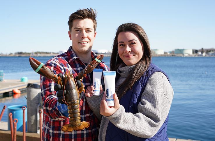 Couple on a dock holding a lobster and skincare tubes labeled MARIN, harbor in background.