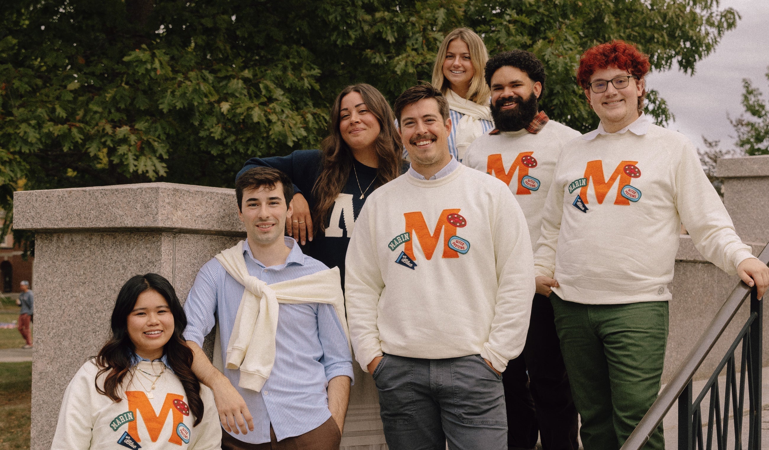 Seven people on campus steps wearing matching sweatshirts with a large orange 'M', smiling and posing outdoors.