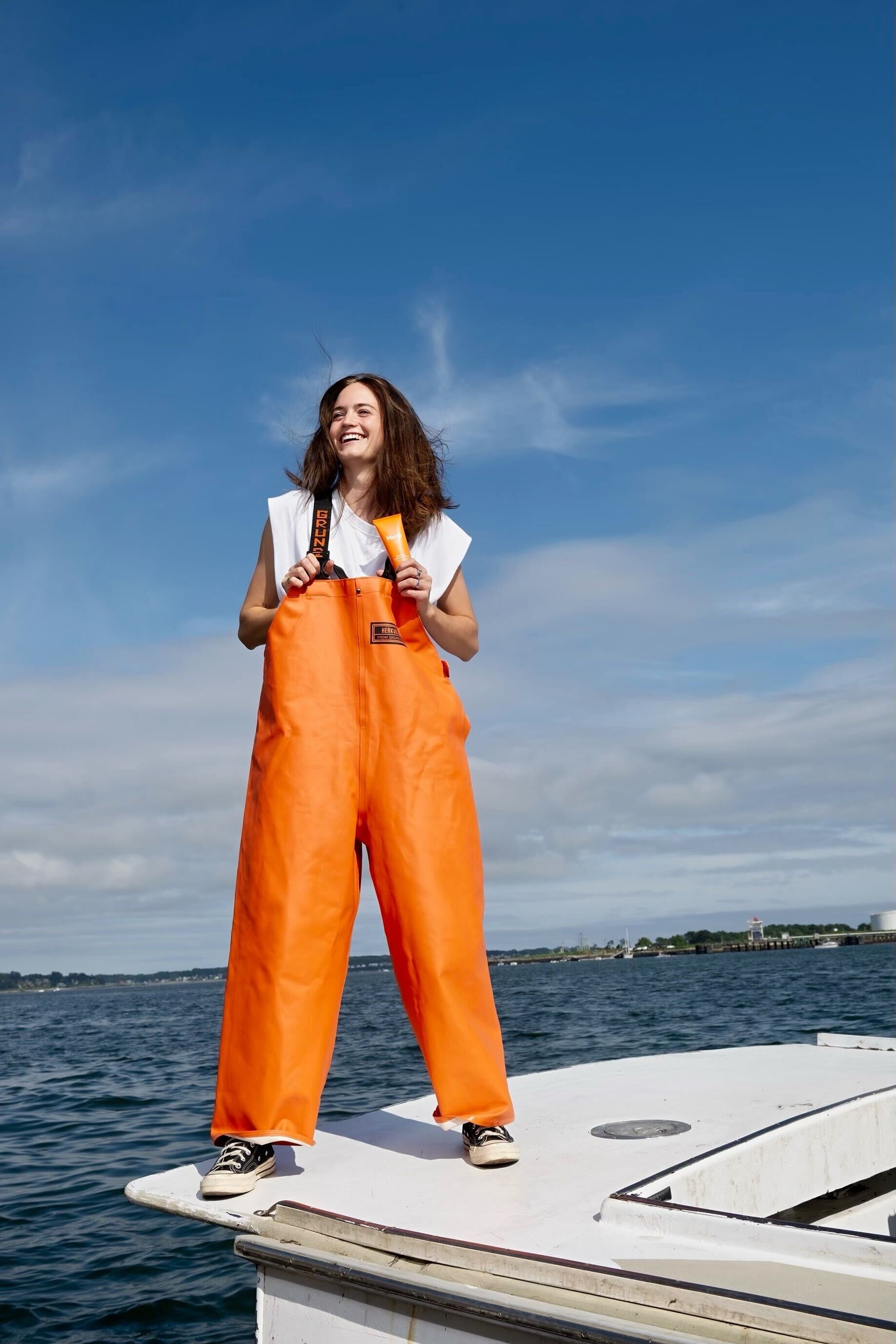 Smiling woman in bright orange fishing overalls stands on a boat against a blue sky, holding a tube of skincare product with the ocean and shoreline in the background.