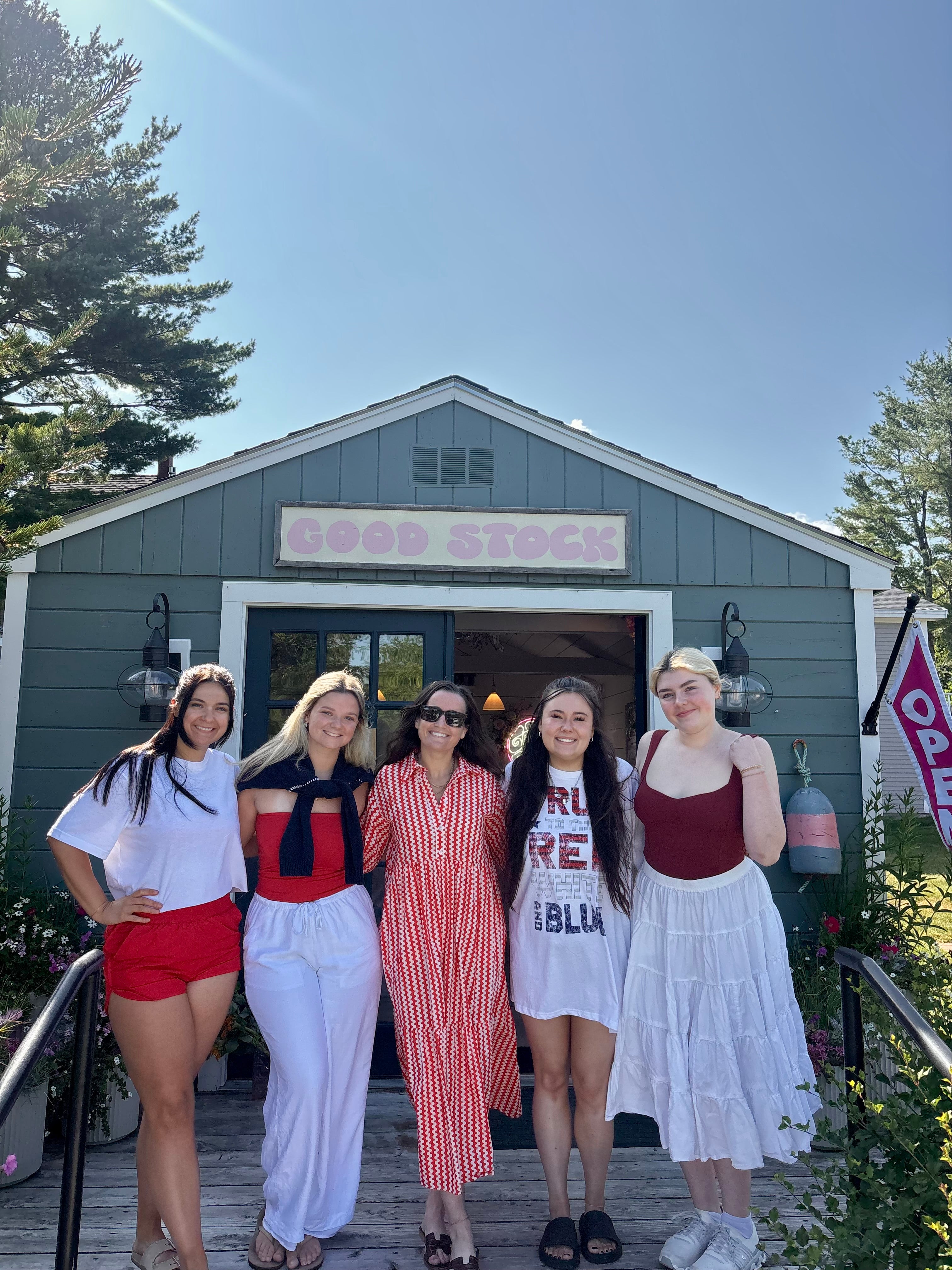 Five women posing on a dock in front of a green shop with sign 'GOOD STOCK' and an 'OPEN' flag