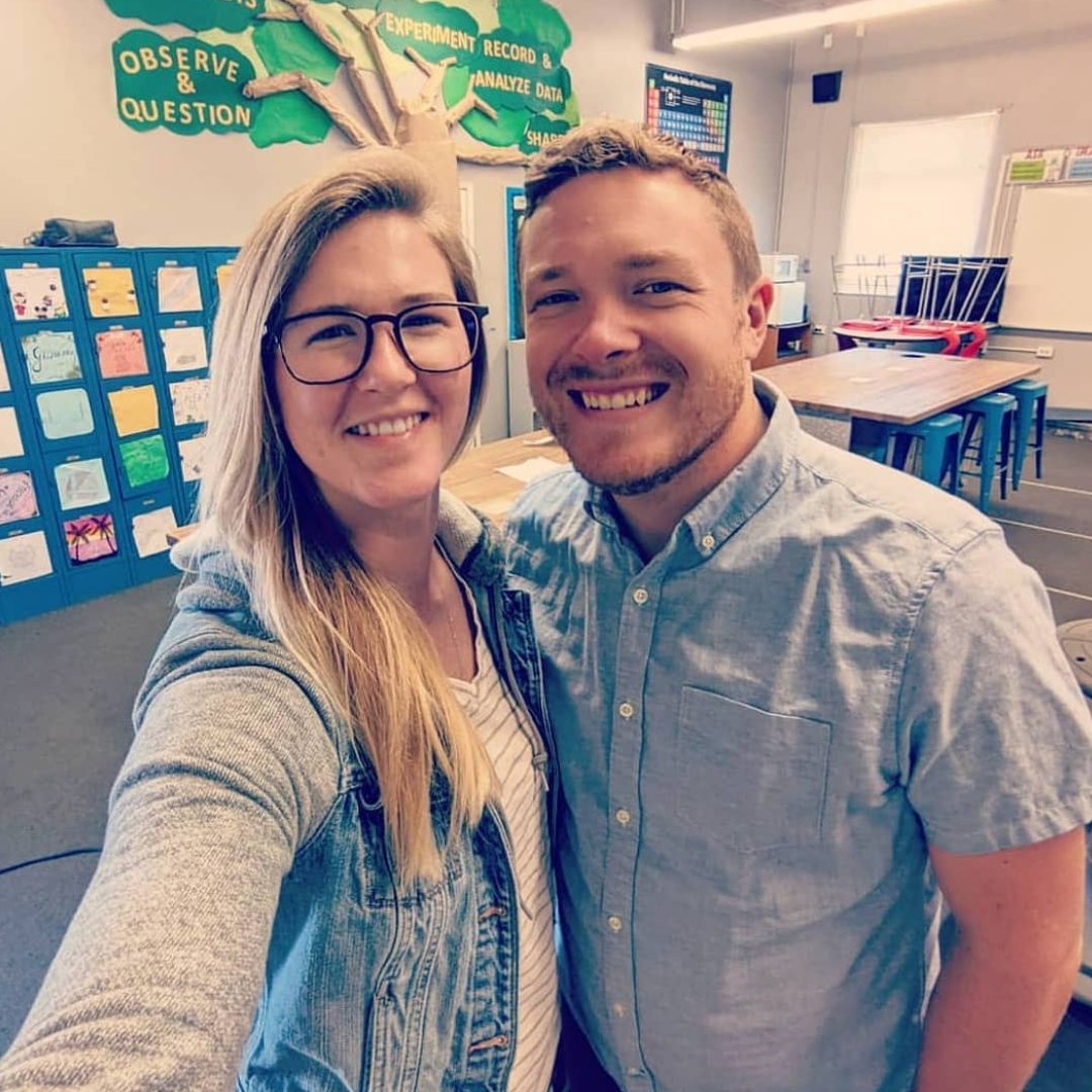 Two smiling adults selfie in a classroom — woman with glasses, blonde hair, denim jacket; man in light-blue button shirt.
