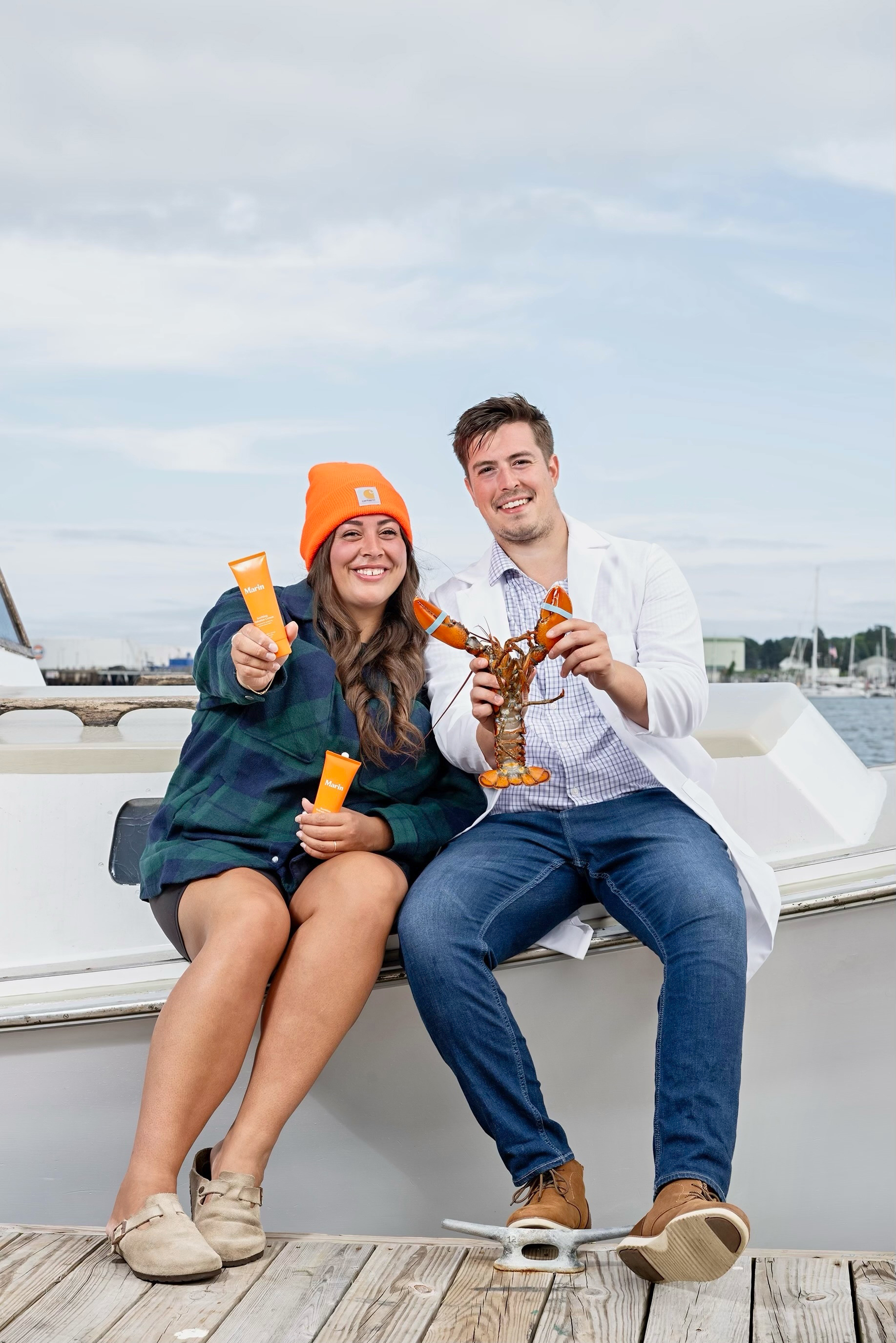 Two smiling people on a dock holding orange Marin Skincare tubes; man holds a live lobster