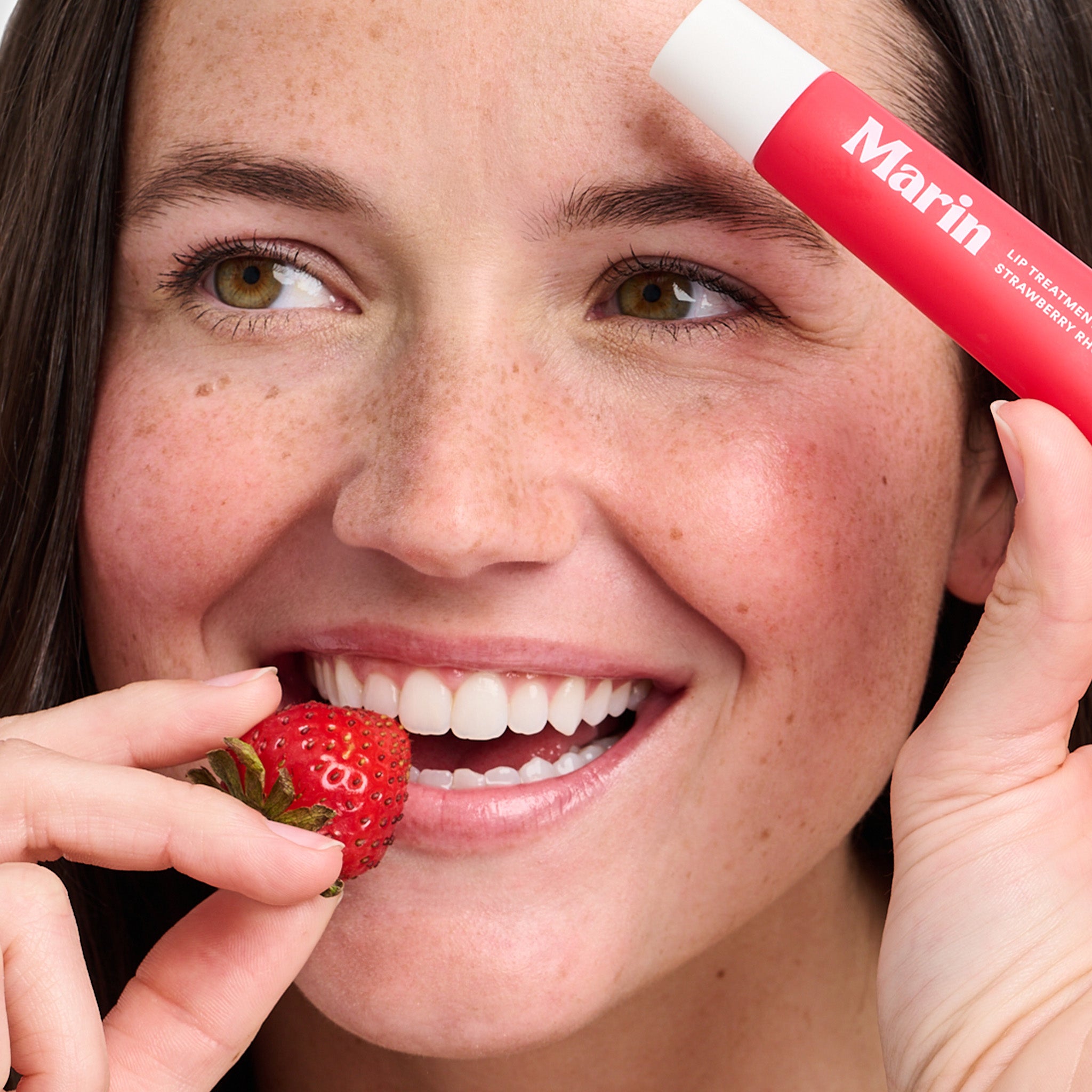 Smiling woman holding a strawberry to her lips and a red lip balm tube labeled Marin LIP TREATMENT STRAWBERRY RHUBARB