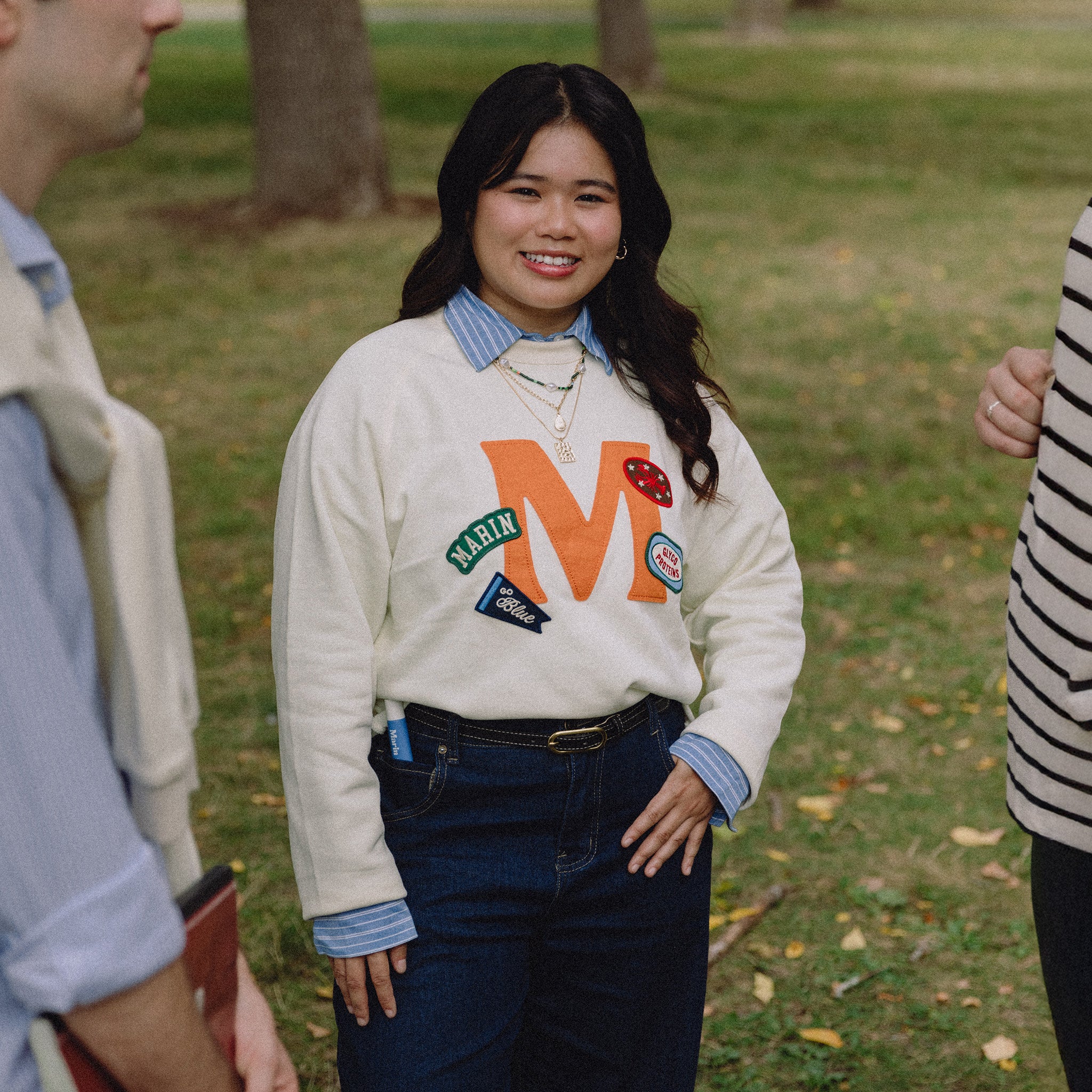 Young woman smiling on grass wearing cream sweatshirt with orange 'M' and patches reading MARIN and go Blue
