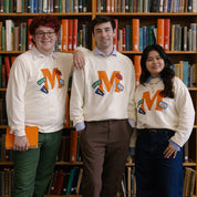Three students wearing cream sweatshirts with large orange M and MARIN patches standing in front of library shelves