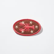 Oval brown embroidered patch with a red lobster motif, six cream stars, and a red border.