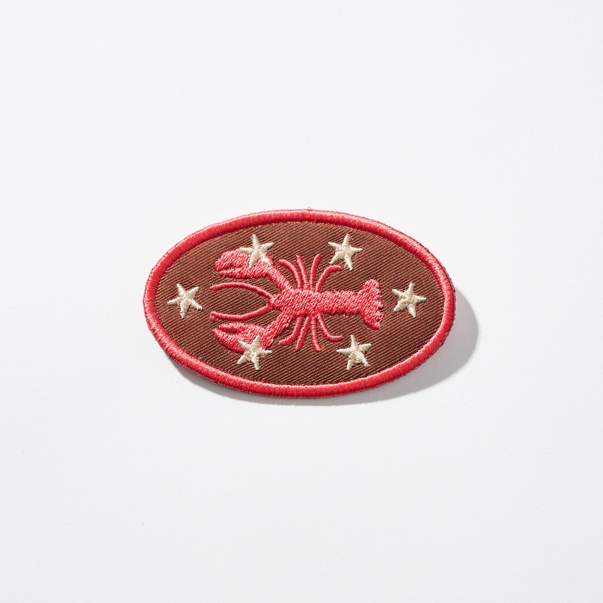 Oval brown embroidered patch with a red lobster motif, six cream stars, and a red border.