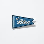 Blue triangular embroidered pennant patch with cream script 'Blue' and two small cream embroidered circles.