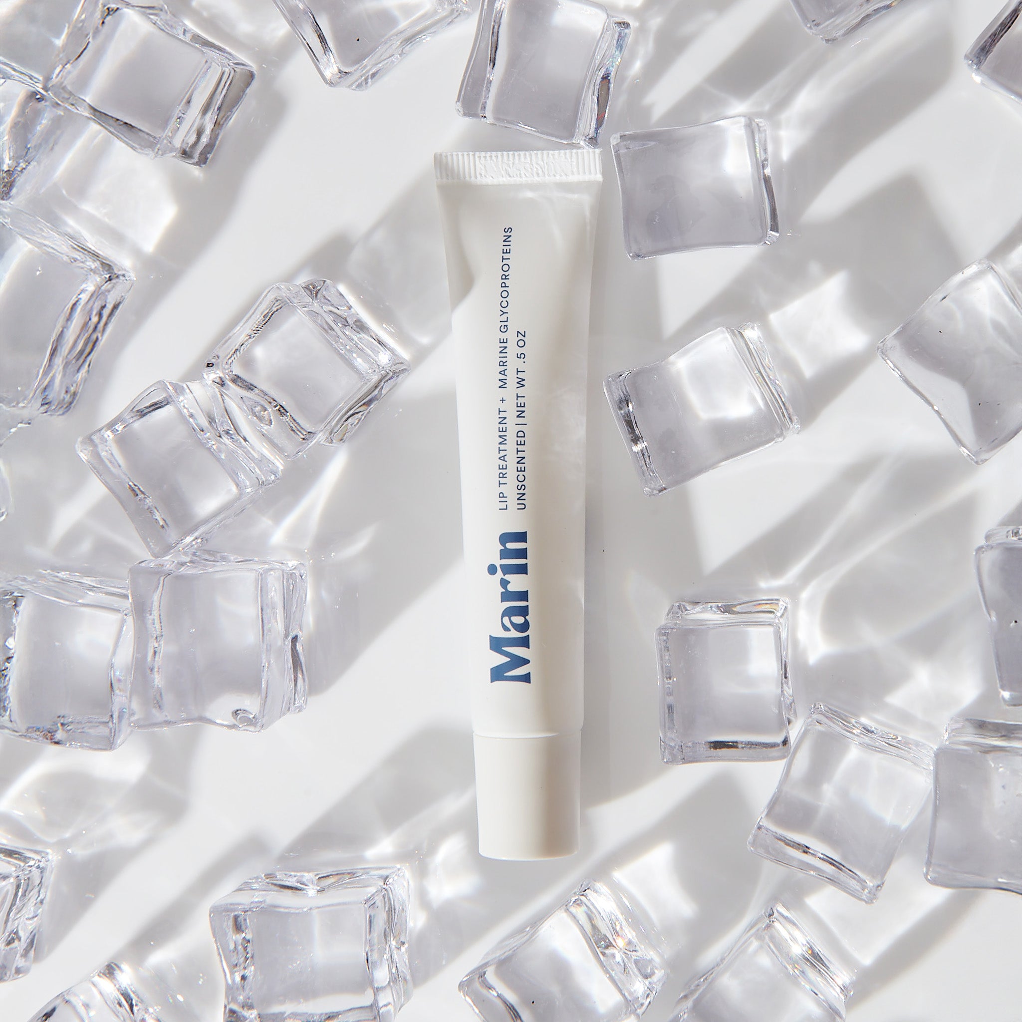 Marin lip treatment tube among clear ice cubes — 'LIP TREATMENT + MARINE GLYCOPROTEINS UNSCENTED NET WT .5 OZ'
