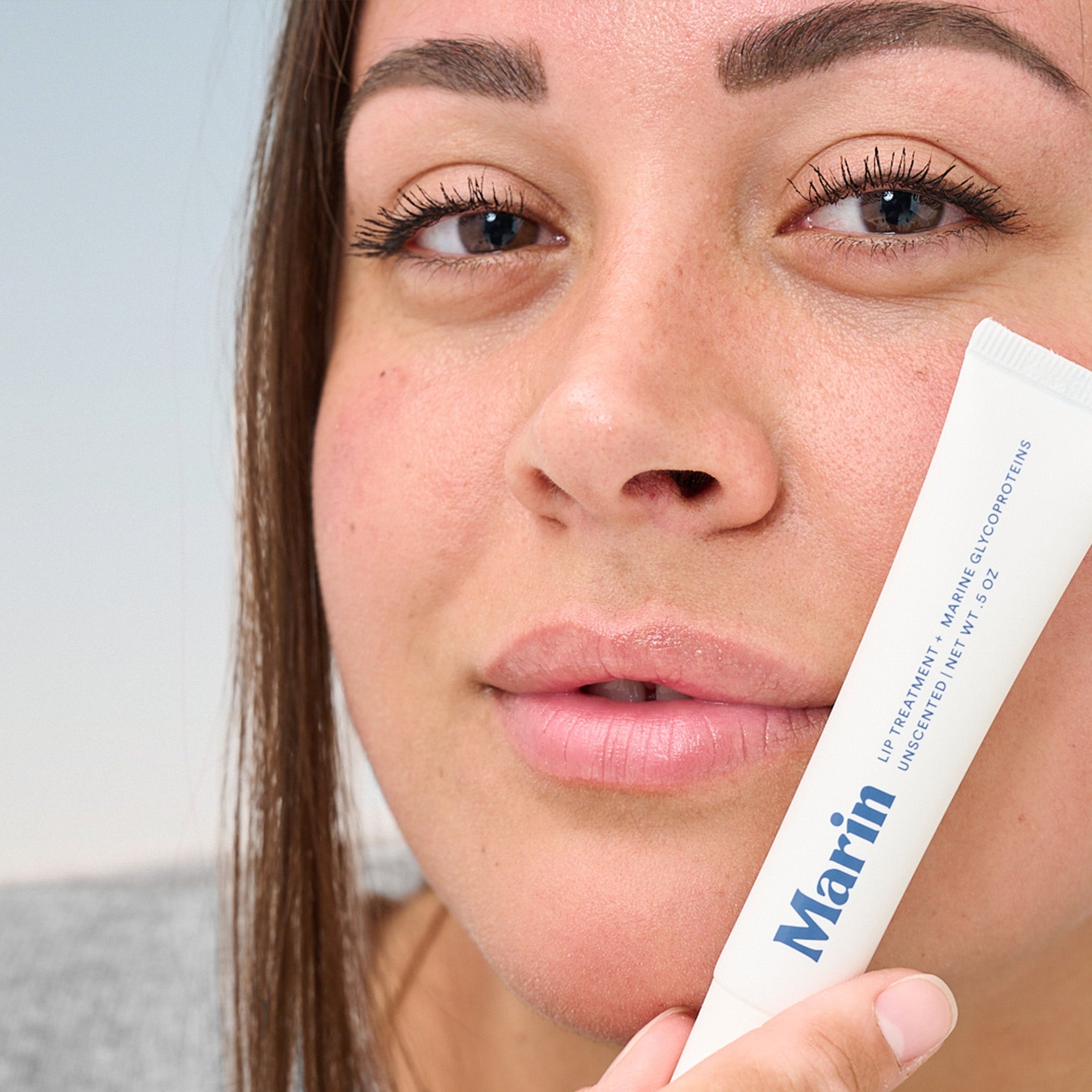 Close-up of woman's lips holding tube reading Marin; LIP TREATMENT + MARINE GLYCOPROTEINS UNSCENTED NET WT .5 OZ