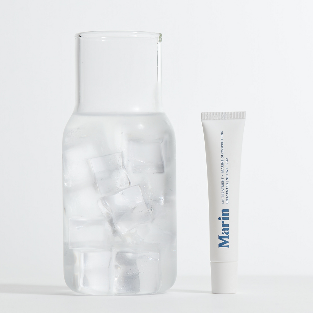 Tube labeled Marin (LIP TREATMENT + MARINE GLYCOPROTEINS; UNSCENTED NET WT .5 OZ) standing next to glass carafe with ice.