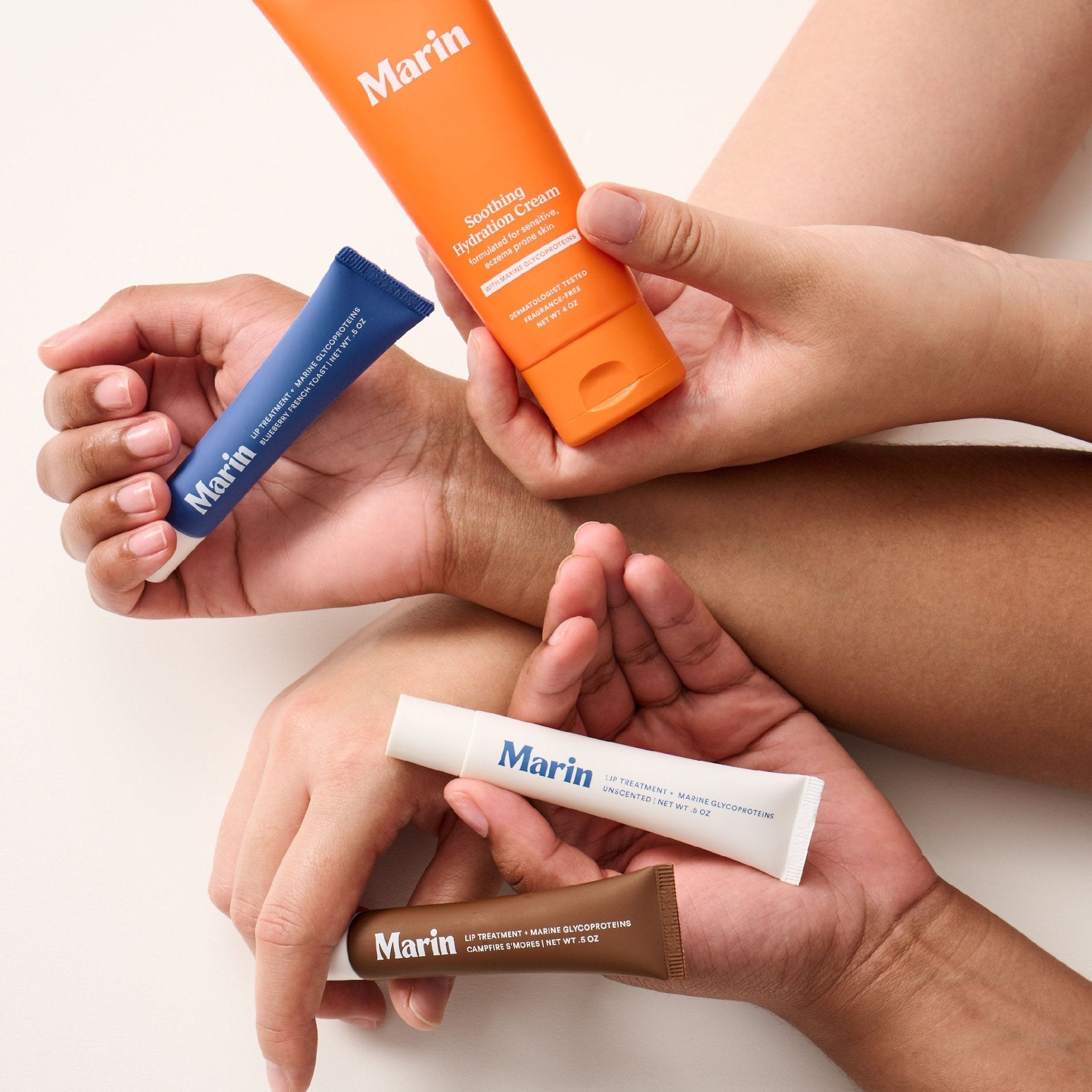 Close-up of several hands holding Marin skincare products, including an orange Soothing Hydration Cream tube and multiple lip treatment tubes in blue, white, and brown.
