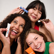 Three smiling women with diverse skin tones holding Marin skincare tubes (white, blue, brown, orange) near their faces.