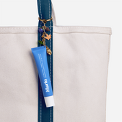 Canvas tote with blue stitched strap and gold charms; Marin lip treatment tube hanging from the strap