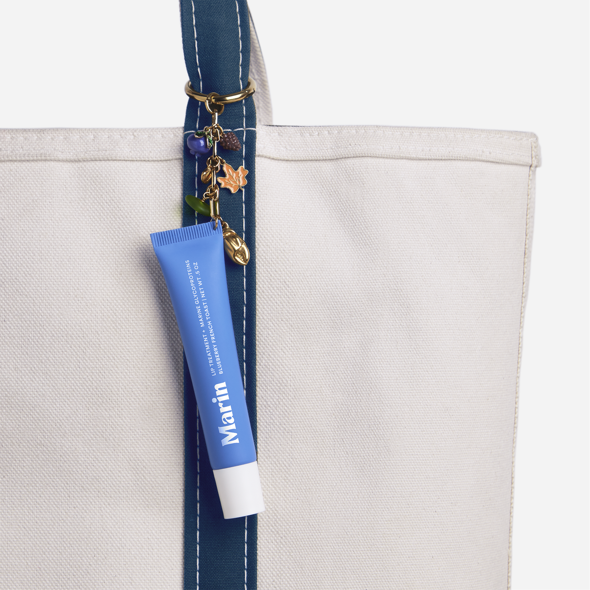 Canvas tote with blue stitched strap and gold charms; Marin lip treatment tube hanging from the strap