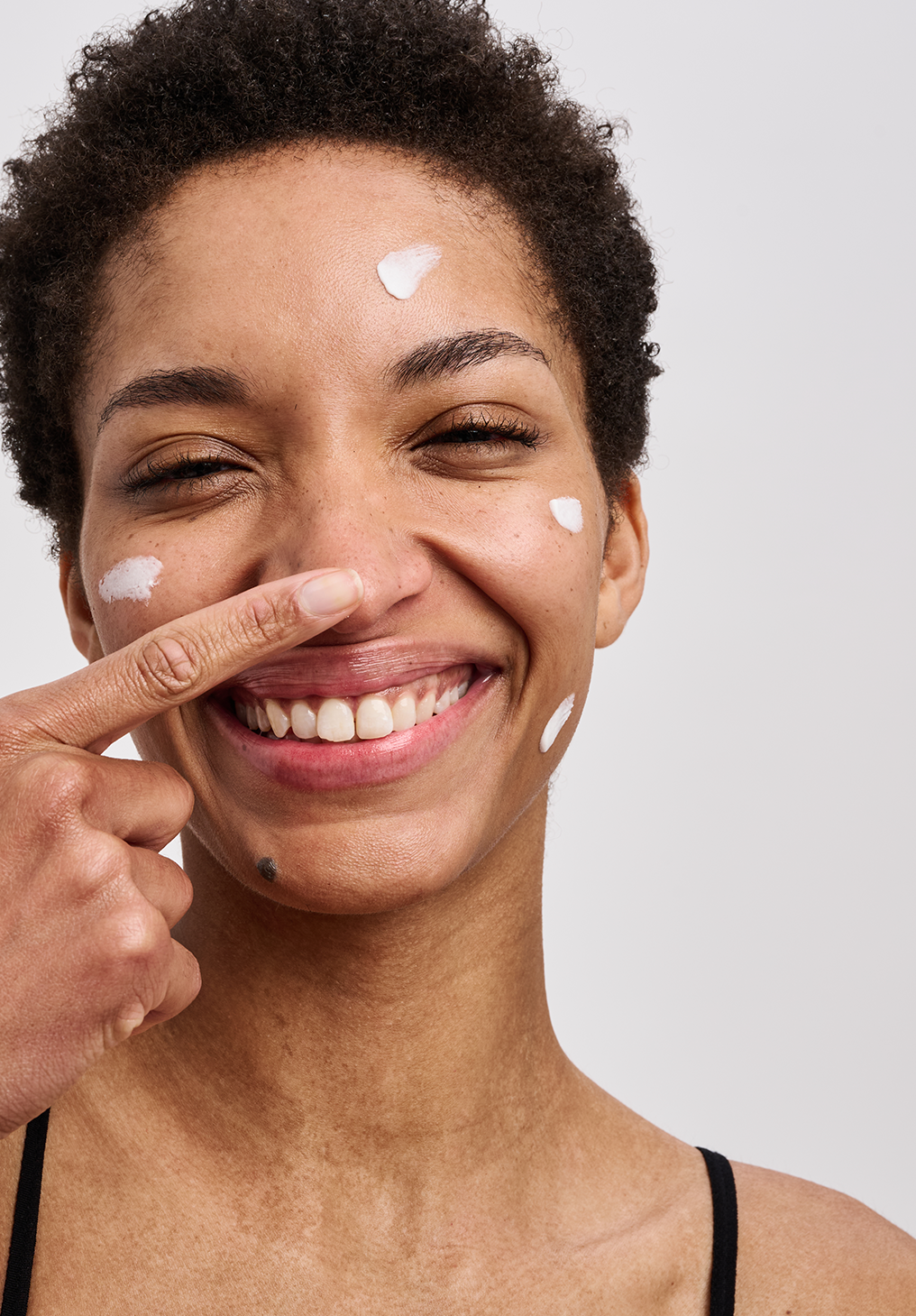 Smiling woman with short curly hair and white moisturizer dots on forehead, cheeks, and chin