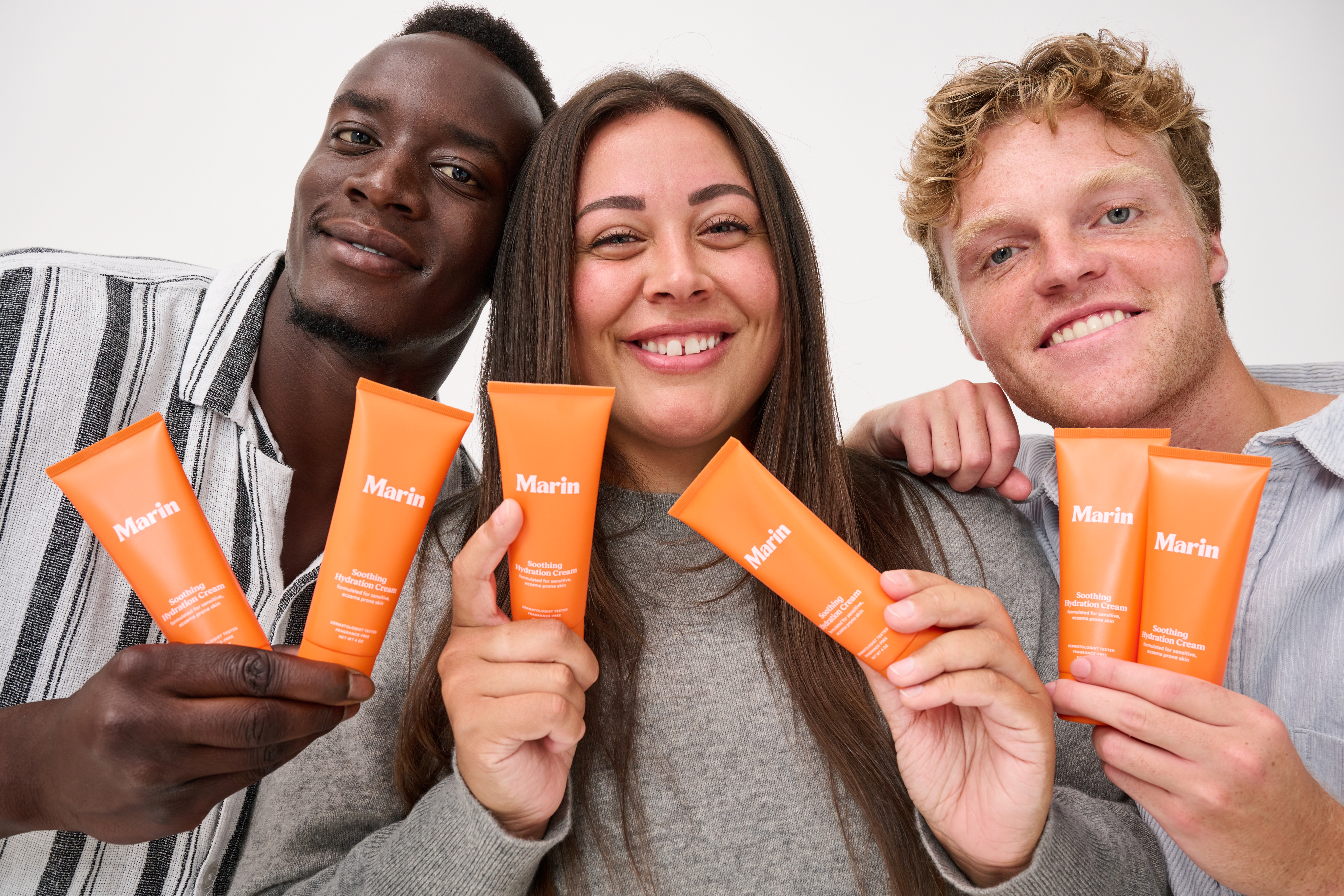 Three smiling people holding orange Marin tubes labeled Marin and Soothing Hydration Cream