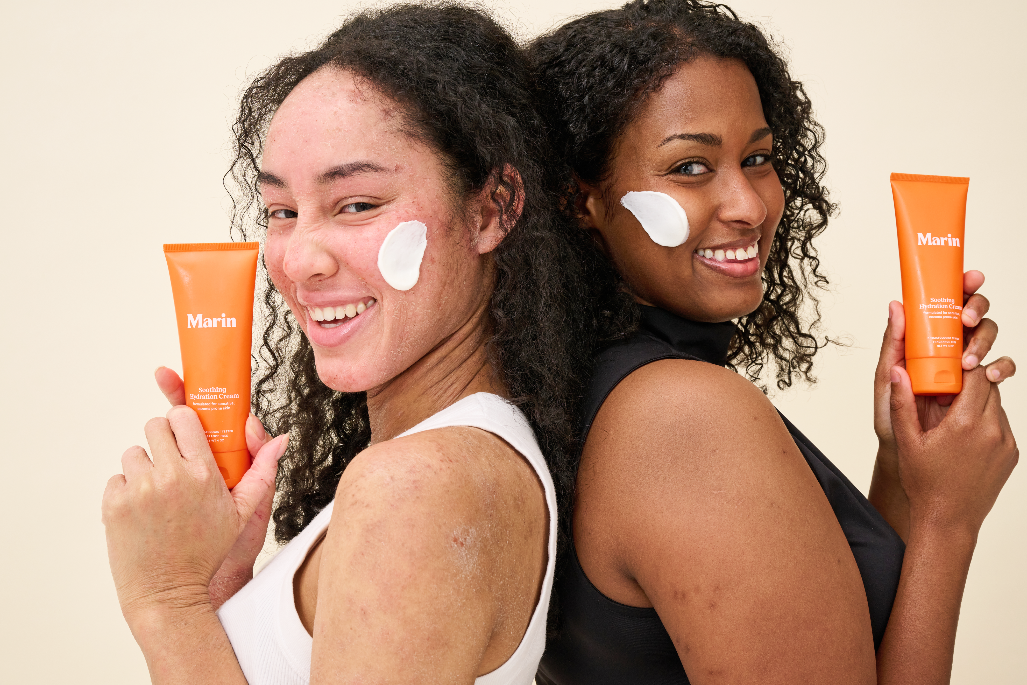 Two smiling women stand back-to-back, each holding an orange tube of Marin Soothing Hydration Cream with a dab of white cream on their cheek against a light background.