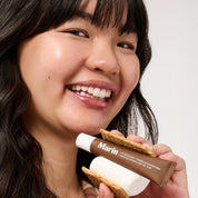 Smiling woman holding Marin (LIP TREATMENT + MARINE GLYCOPROTEINS; CAMPFIRE S'MORES; NET WT .5 OZ) tube as part of a s'more
