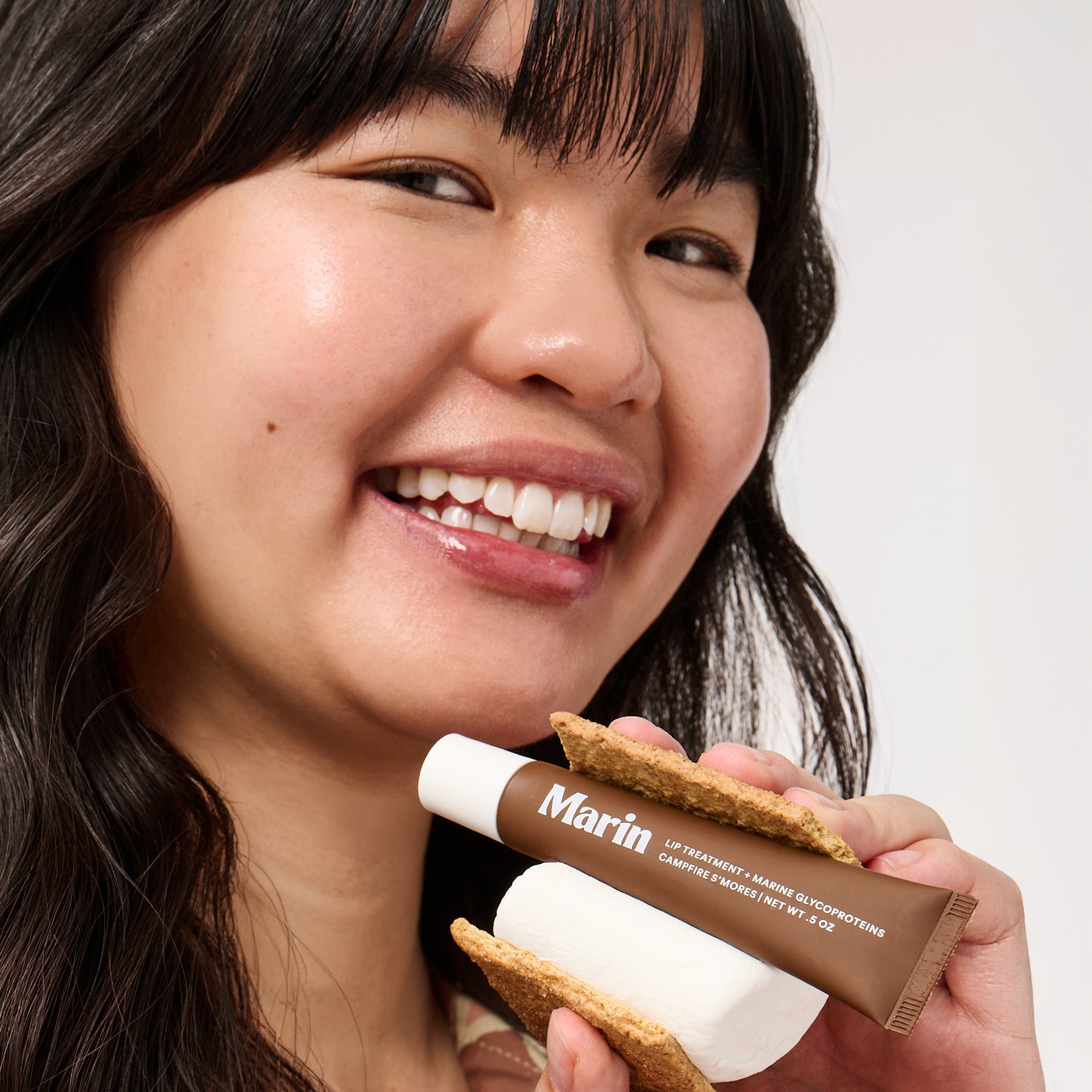 Smiling woman holding Marin (LIP TREATMENT + MARINE GLYCOPROTEINS; CAMPFIRE S'MORES; NET WT .5 OZ) tube as part of a s'more