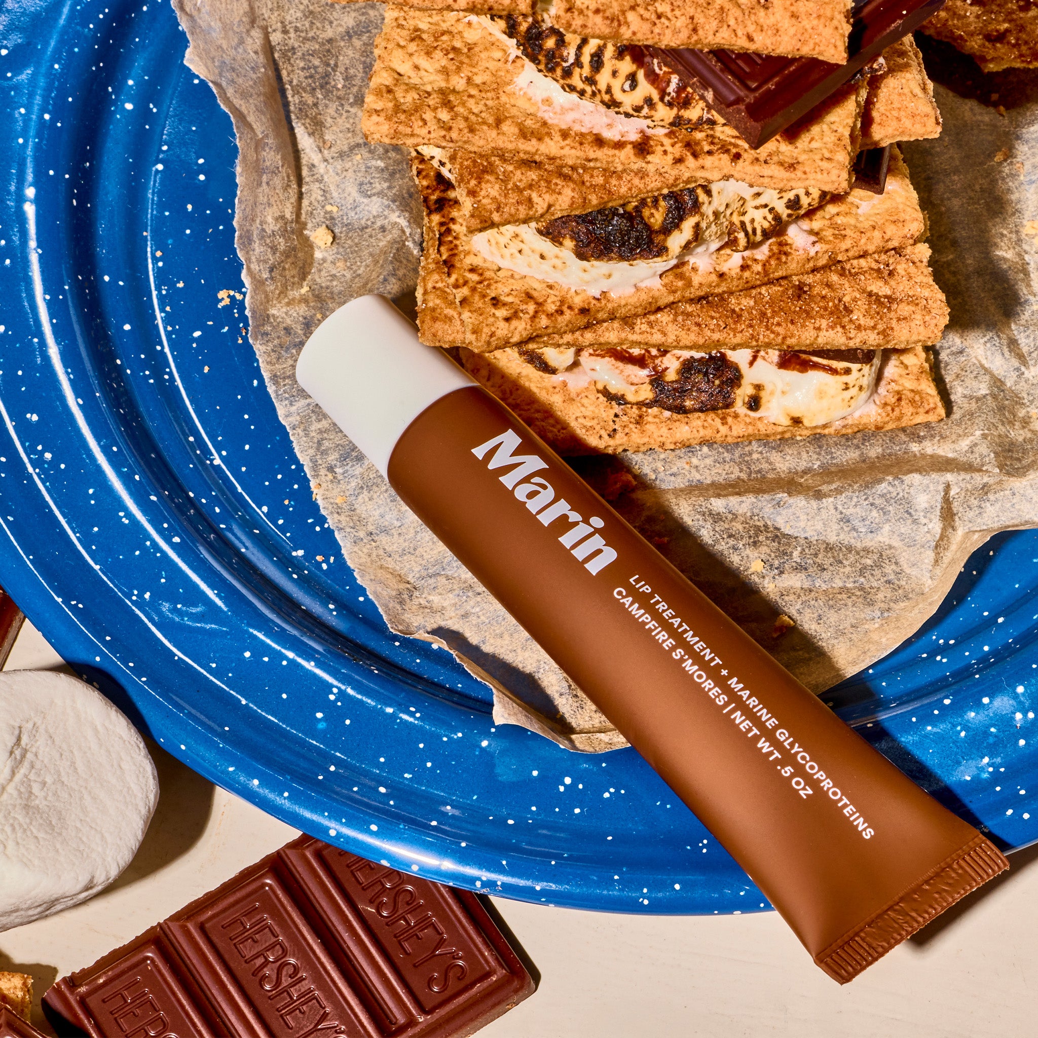 Marin tube labeled LIP TREATMENT + MARINE GLYCOPROTEINS CAMPFIRE S'MORES NET WT .5 OZ on blue plate with HERSHEY'S chocolate.